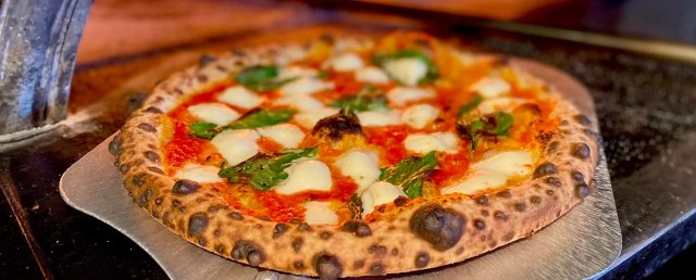 Freshly baked brick-oven pizza topped with melted mozzarella, basil, and rich tomato sauce, resting on a pizza peel inside a wood-fired oven at Square Peg Pizza at east Hartford Golf Course.